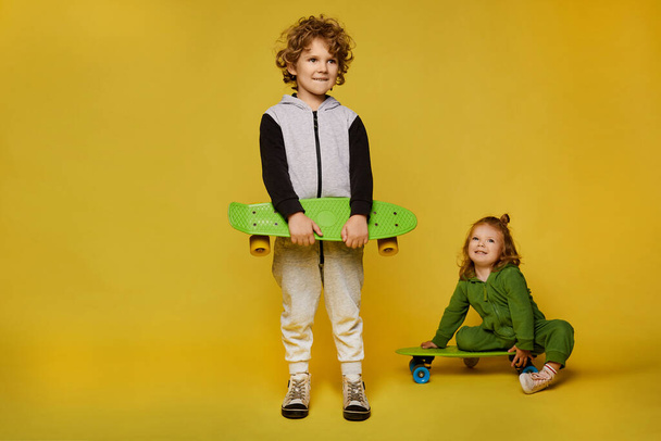 Stylish children in modish outfits posing with skateboards. Little girl and little boy isolated at the yellow background with copy space. Children fashion - Photo, Image