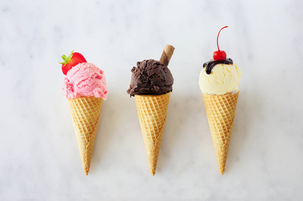 Ice cream cone assortment. Strawberry, chocolate and vanilla in waffle cones. Top view over a white marble background. - 写真・画像