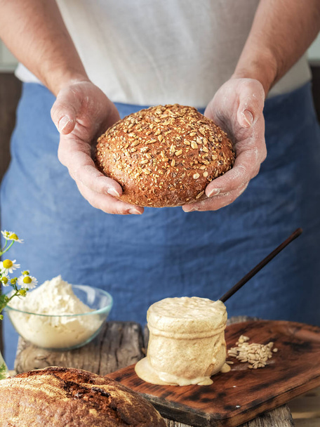 Baker házi készítésű bio kenyeret tart frissen a sütőből, lisztből, élesztőből, sóból és vízből. Házi sütés az elszigeteltség időszakában, kovászos tészta, liszt fából készült asztalon, közelkép - Fotó, kép