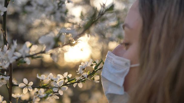 girl removes medical mask and sniffs blossoming tree of apple or cherry at sunset close-up - Photo, Image