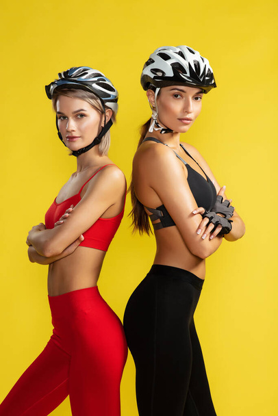 Female cyclists. Two young attractive women in sportswear and biking helmets looking at camera while standing against yellow background. Studio shot - 写真・画像