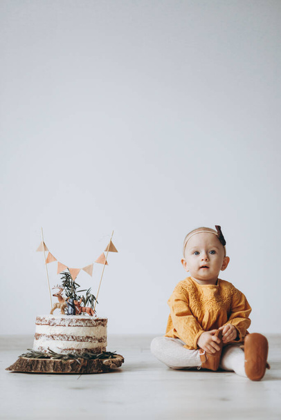 cute little girl dressed in a yellow dress and a bandage sits on the floor next to a holiday cake on a white background - Φωτογραφία, εικόνα