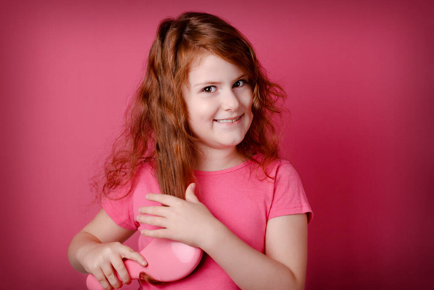 Rotschopf lockiges Mädchen pflegt und kämmt ihre Haare. Kleine Teenie-Mädchen lächeln auf rosa Hintergrund.  - Foto, Bild