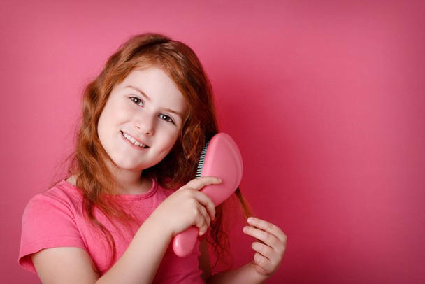 Pelirroja chica rizada es aseo y peinándose el pelo. Niña adolescente sonriendo sobre fondo rosa. Copiar espacio
. - Foto, Imagen