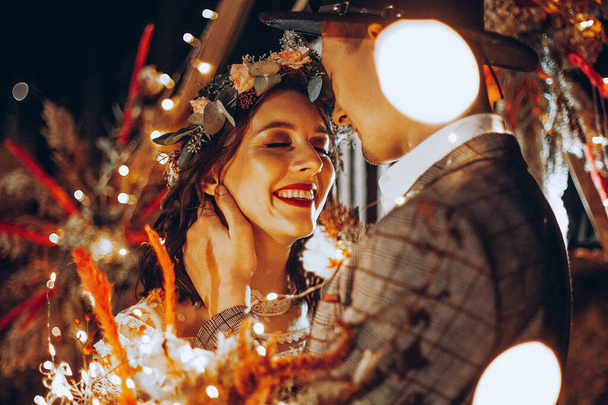 Stylish couple of brides, pretty woman in wedding dress and young man in fedora hat. Romantic wedding ceremony in nature. The lights of the electric garland illuminate the wedding party. - Valokuva, kuva
