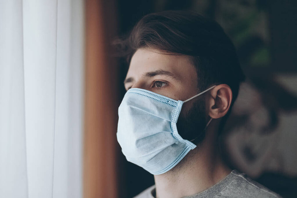 portrait of a young man wearing a medical mask and quarantined at home or in a hospital ward opposite a window - Fotoğraf, Görsel