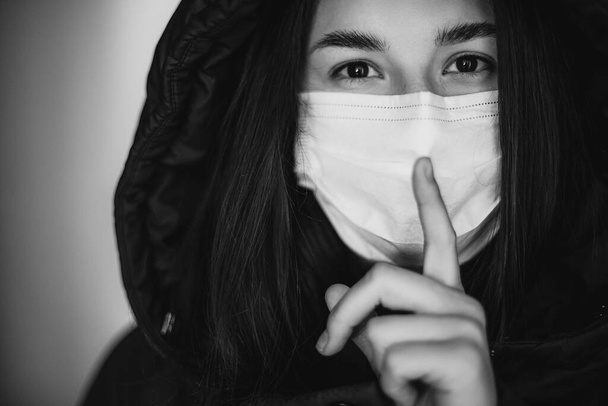 a young woman wearing a medical mask shows a hand sign that means it's a secret and needs to be silent. Medical concept of spread of viral diseases and quarantine - Foto, imagen