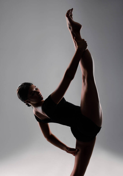 A flexible young female gymnast in sportswear performs a stretch. Training, gymnastics, acrobatics on a gray background. Sports motivation, physical education - Foto, Bild