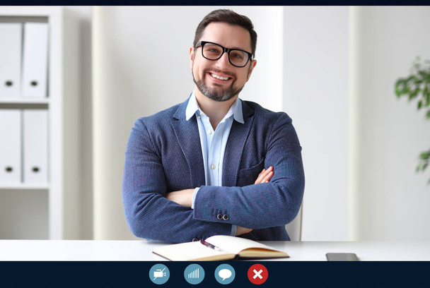 Zelfverzekerde zakenman met gekruiste armen glimlachend en kijkend naar de camera terwijl hij op kantoor zit en online bijeenkomsten bijwoont - Foto, afbeelding