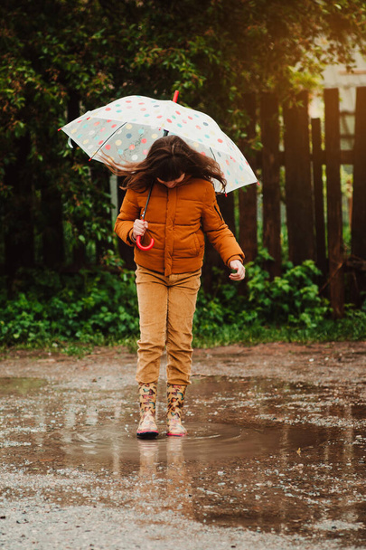 Szczęśliwa dziewczynka z parasolem i gumowymi butami skacząca w kałuży. Zimny wiosenny deszczowy dzień. Dziecko pod parasolem - Zdjęcie, obraz