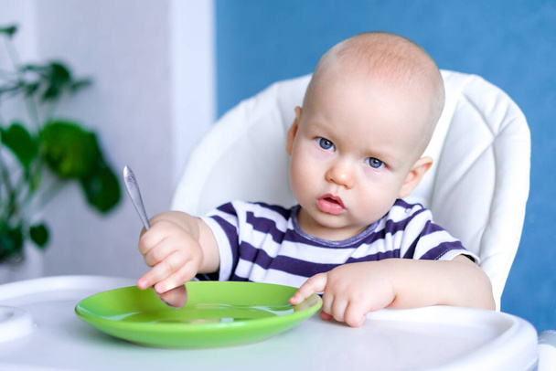 baby food. little serious caucasian child with spoonand empty green plate on highchair. Newborn infant kids - Фото, зображення