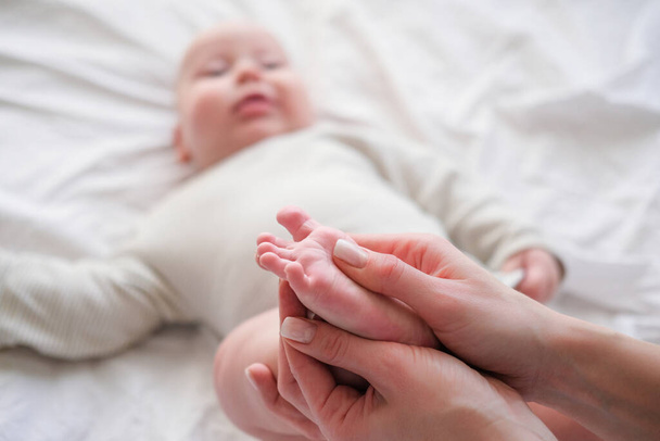 Baby feet in mother hands. Young caucasian woman makes massage for happy infant baby on white bed at home. Babycare, sport and happy motherhood. - Foto, imagen