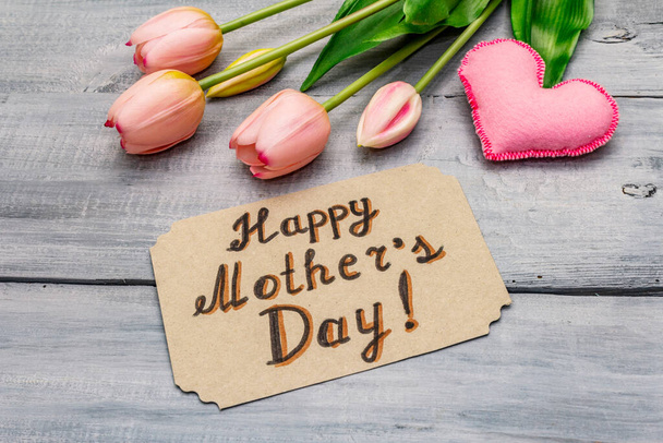 Üdvözlőlap a Boldog Anyák Napjára. Gyengéd rózsaszín tulipán, kézzel készített filc szív, nulla hulladék csomagolási ajándék. Fa táblák háttér - Fotó, kép