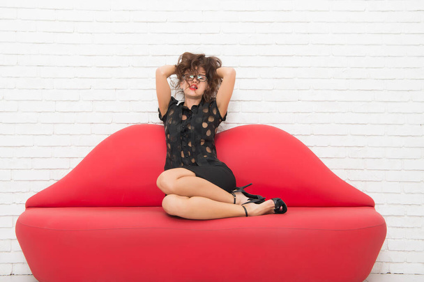 relaxed girl sitting on red sofa. sexy model on leather couch. beautiful luxurious woman seducing you. feel the desire. beauty and fashion concept. portrait of beautiful sensual woman - 写真・画像
