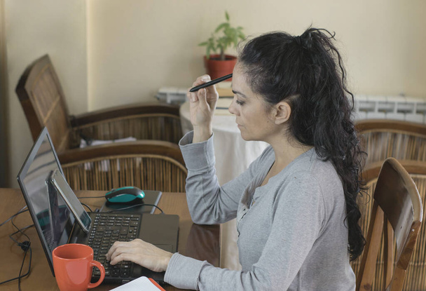 Brünette Frau, die zu Hause telefoniert und nachdenklich auf ihren Computerbildschirm blickt - Foto, Bild