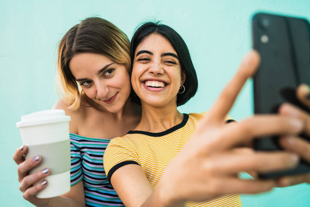Portrait of lovely lesbian couple having fun and taking a selfie with mobile phone against light blue background. LGBT concept. - Fotoğraf, Görsel
