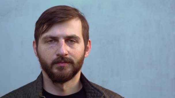Handsome young Caucasian man with thick heavy beard, freckles in black jacket with brown hair looks directly at camera on gray wall background. Portrait of serious guy outdoor on sunny day close up. - Footage, Video