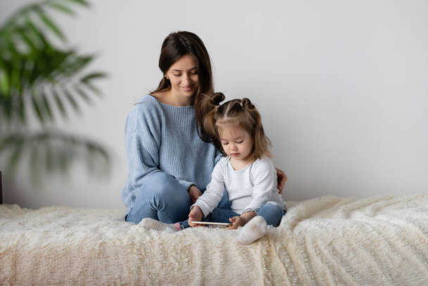 Egy két éves kislány ül az ágyon az anyjával. Játssz mobilon. Okostelefon egy gyerek kezében. Fehér fal hely szöveges. - Fotó, kép