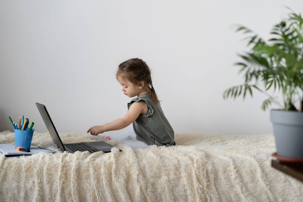 La petite fille reste assise à la maison sur le lit pendant deux ans. Regarder un film sur un ordinateur portable. Mur blanc, endroit pour le texte
. - Photo, image