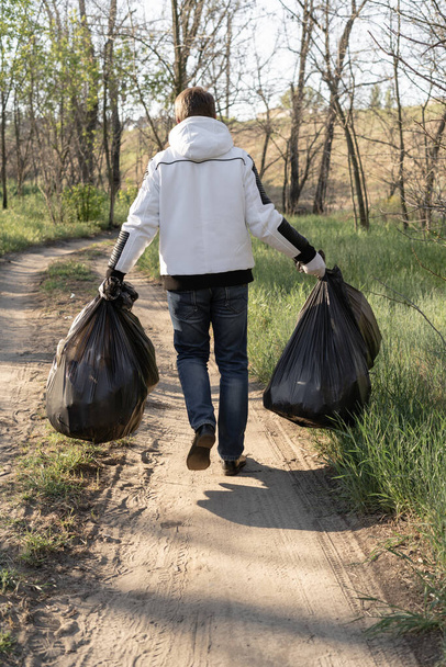 Концепция праздника Дня Земли. Молодой человек в джинсах выносит три полных пакета мусора из парка. Эко-волонтеры убирают лес
. - Фото, изображение