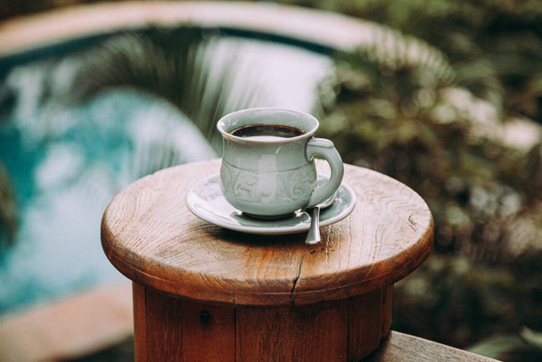 Cup of coffee on wooden table at pool in tropical garden. - Фото, зображення