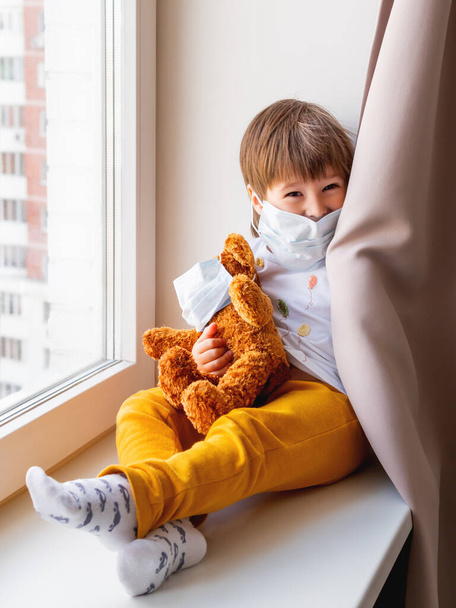 Toddler boy sits with teddy bear in medical masks. Kid with plush toy plays hide-and-seek behind curtains. Browh plush bear and child on home quarantine because of coronavirus COVID-19. - Фото, изображение