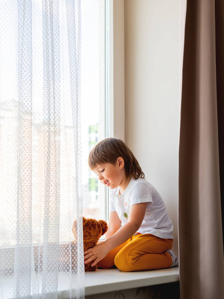 Toddler boy sits with teddy bear. Kid with plush toy look through window outside. Brown bear and child on home quarantine because of coronavirus COVID-19 waiting for somebody. - Foto, Bild