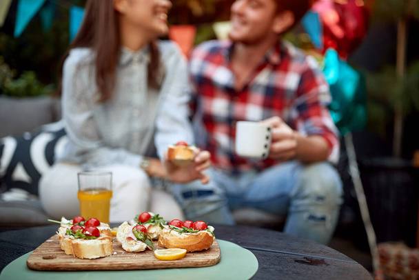 focus on sandwiches with cherry tomato and orange juice with couple in the background out oif focus talking, smiling. People, food and beverage - Photo, Image