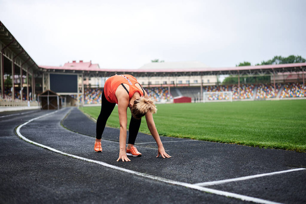 Retrato de larga duración de una mujer rubia, lista para correr en un estadio en verano, deportista en forma, con leggings negros y top naranja y zapatillas de deporte, advirtiéndose antes del entrenamiento
. - Foto, imagen