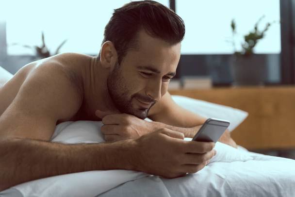 Cheerful young guy using mobile phone in bed - Photo, Image