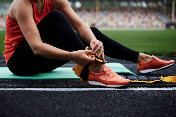 Close-up picture of woman's legs, showing process of putting on yellow ankle weights. Young woman, wearing black leggings and orange sneakers, is preparing herself for fitness training with weights.on - Fotografie, Obrázek