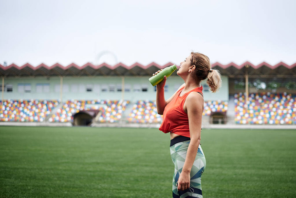 Young fit blond woman, wearing orange top and green leggings, drinking water from light green bottle, Refresh yourself during sport exercise on stadium with green grass. Athlete on training - Φωτογραφία, εικόνα