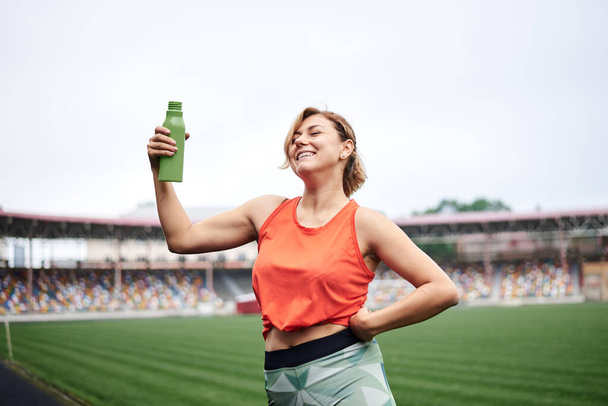 Young fit blond woman, wearing orange top and green leggings, drinking savoring water from light green bottle, smiling. Refresh during sport exercise on stadium with green grass. Athlete on training - Фото, изображение
