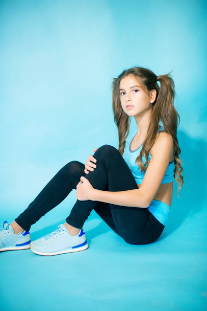 Cute teenager girl with long blond hair in fitness clothes on a blue background in the studio. Sports fashion. Sport for youth. - Фото, изображение
