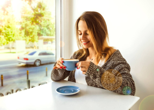 beautiful young woman drinks coffee sitting by the window. Beautiful teenager sitting by the window having a hot drink. - Фото, зображення