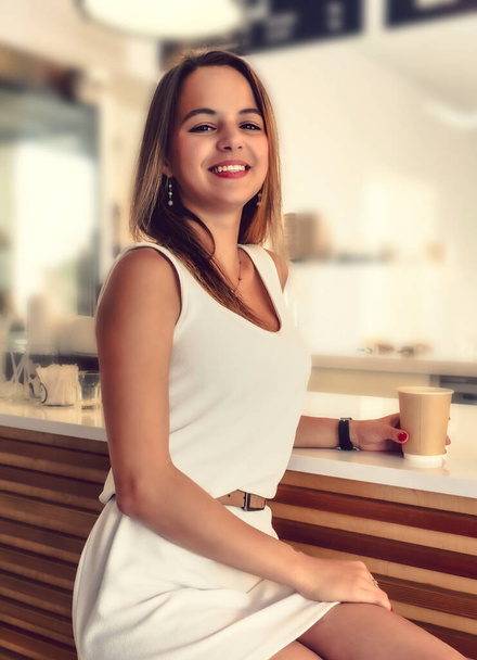 Young beautiful girl holding a cup of coffee in cafe. Outdoor portrait of young beautiful woman posing while traveling holiday vocation. Girl with coffee. Female traveler, travel vacation fun summer. - 写真・画像