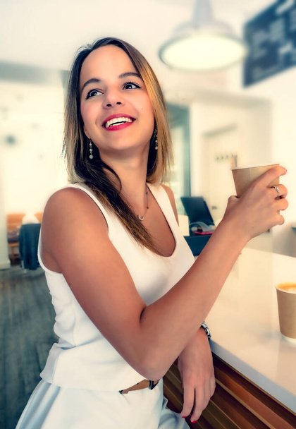 Young beautiful girl holding a cup of coffee in cafe. Outdoor portrait of young beautiful woman posing while traveling holiday vocation. Girl with coffee. Female traveler, travel vacation fun summer. - Фото, зображення