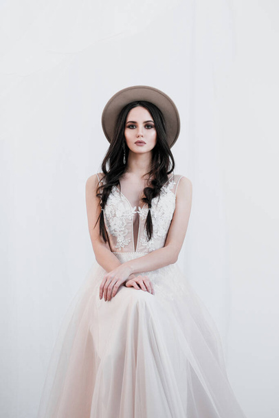 Portrait of bride. A young attractive brunette woman with long black hair in braids. White wedding dress and beige hat. Rustic style, bouquet of wheat spikes. - Fotó, kép