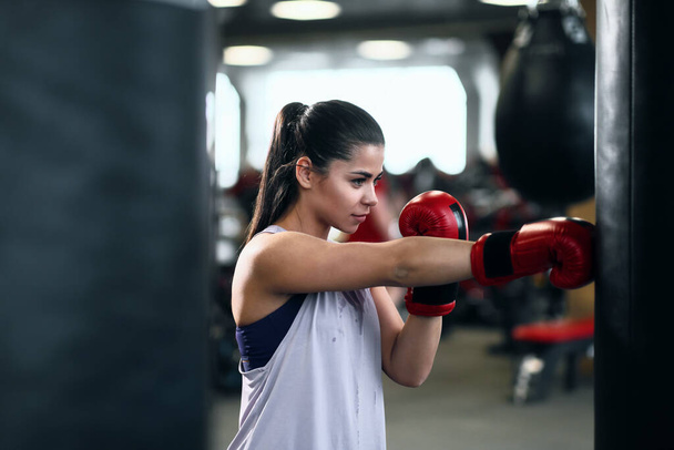 Sportovní mladá brunetka žena ve sportovním oblečení a červené boxerské rukavice vlaky naráží na boxovací pytel ve fitness tělocvičně. - Fotografie, Obrázek