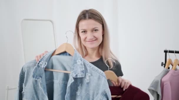 A young attractive millennial woman in white room tries on and chooses clothes. Looks in mirror and compares outfits. Wardrobe room of house or fitting room in store. - Metraje, vídeo