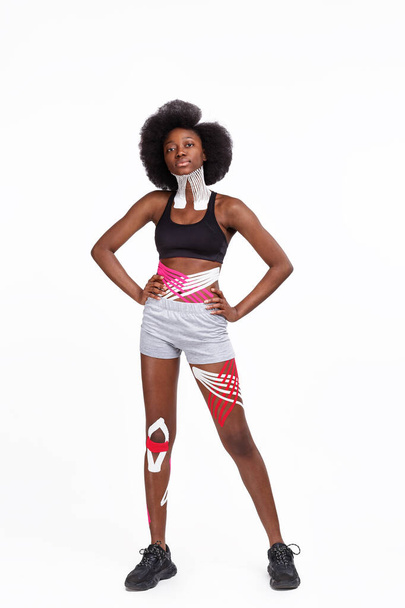 Young smiling female African American athlete on white background.Kinesiology taping.Kinesiology tape on patient neck, tummy, hip and knee.Sport physical therapy,recovery concept,alternative medicine - Valokuva, kuva