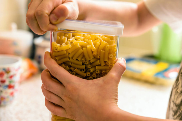 childs hands opening macaroni pasta inside clear storage container on kitchen worktop - Foto, imagen