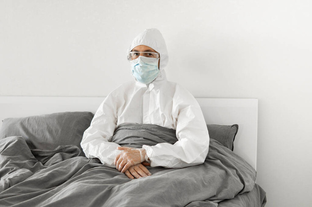 Portrait of man in protective white suit and face mask sitting in his bed in his bedroom because of covid-2019 and wait for a end of coronavirus quarantine. Self isolation. - Photo, Image