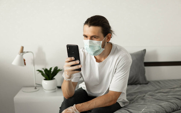 Man in medical mask is using a phone at his home sitting on a bed because of coronavirus epidemic. Remote work during pandemic. Stay home during COVID-19 quarantine concept. - Photo, Image