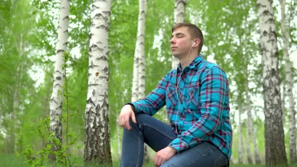 Молодий чоловік сидить у природі з навушниками у вухах. Чоловік сидить на пні в березовому лісі і слухає музику
. - Кадри, відео
