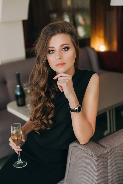 Romantic sad woman with long hair holds glass of champagne. Gorgeous long healthy hair. Brown-hairedmodel with waving hair in black. Professional makeup, sexy sensual lips - Photo, image