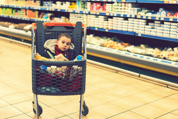 Όμορφο μωρό στο καλάθι αγορών - τρόλεϊ. Ένα τρόλεϊ με προϊόντα στα οποία κάθεται το παιδί. Πλήρες καλάθι με φαγητό στο σούπερ μάρκετ. Στο καρότσι κάθεται ένα μωρό. τονισμένο - Φωτογραφία, εικόνα