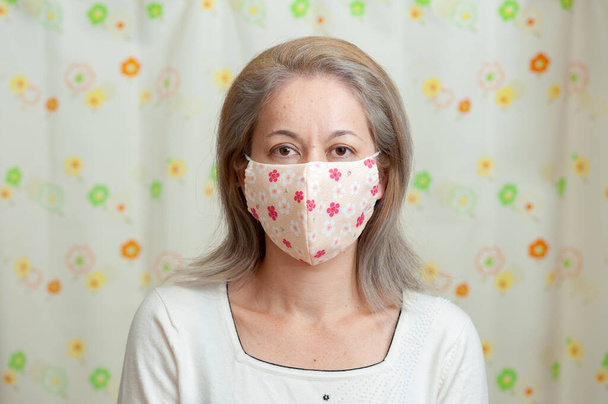 Vorderseite Porträt einer Mischlingsfrau mittleren Alters mit DIY-Gesichtsmaske aus Stoff zum Schutz vor Coronavirus (COVID-19) und anderen Infektionskrankheiten. Floraler Vorhang Hintergrund. - Foto, Bild