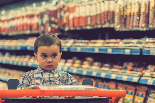 Uroczy chłopiec ubrany w koszulę w kratę w supermarkecie w wózku. Na tle półek z produktami mięsnymi i kiełbasami. stonowana - Zdjęcie, obraz