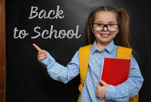 Cute little child wearing glasses near chalkboard with phrase BACK TO SCHOOL - Fotó, kép